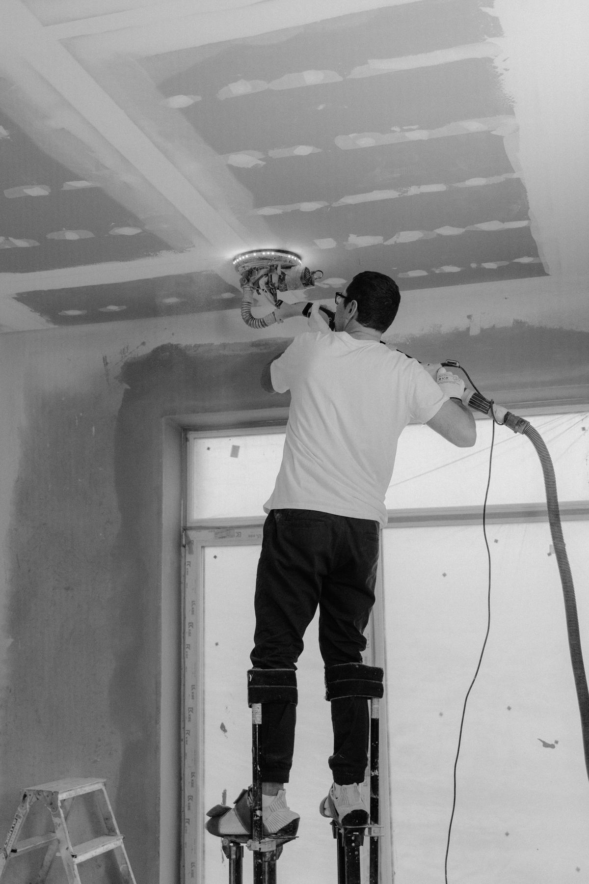 Tradesman on drywall stilts running a ceiling sander across taped seams, shown in black and white