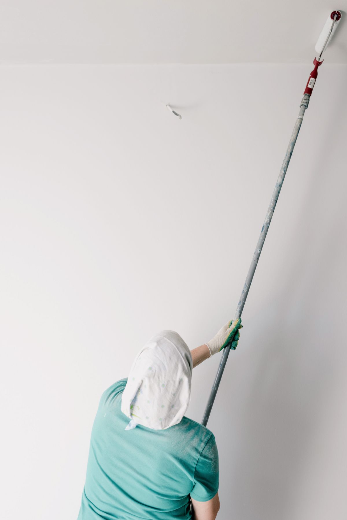 Painter in a headscarf and gloves rolling white paint onto a ceiling with an extension pole